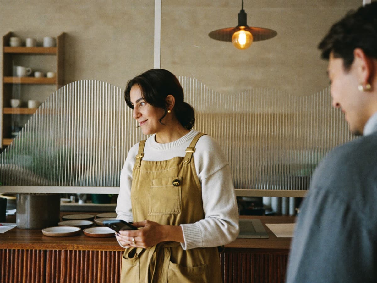 Dueña de una cafetería platica con un cliente en la barra mientras su asistente responde los mensajes